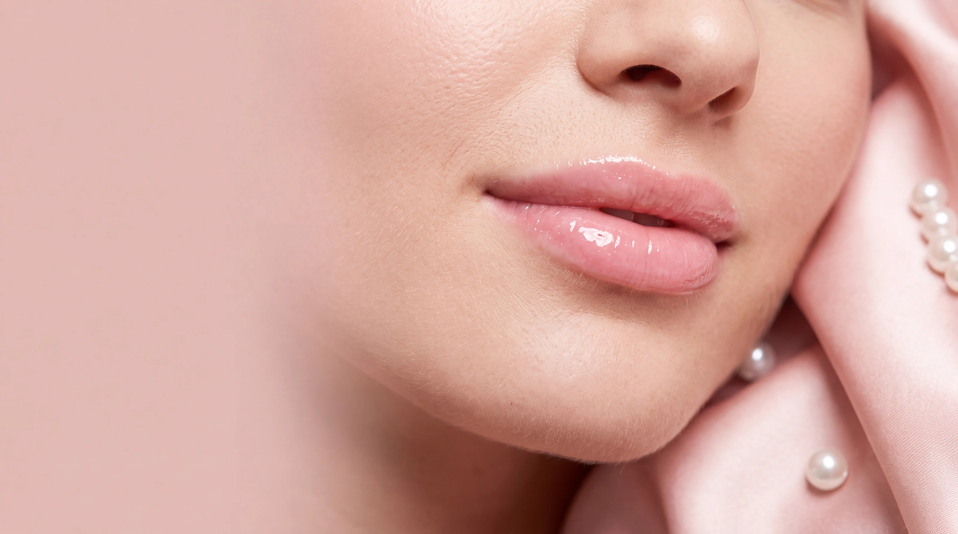 Close-up of glossy pink lips against blush satin and pearls
